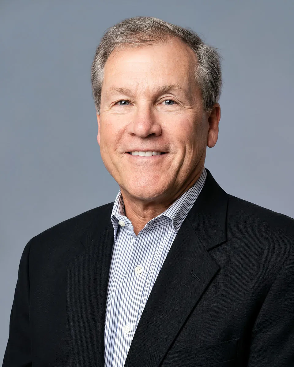 A professional remote headshot of a man in a dark blazer, captured with a phone.