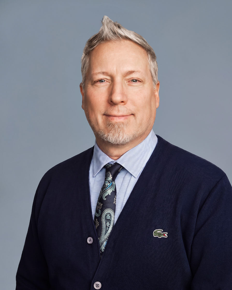 Formal company website headshot of a man in a dark suit with a clean blue-gray background.