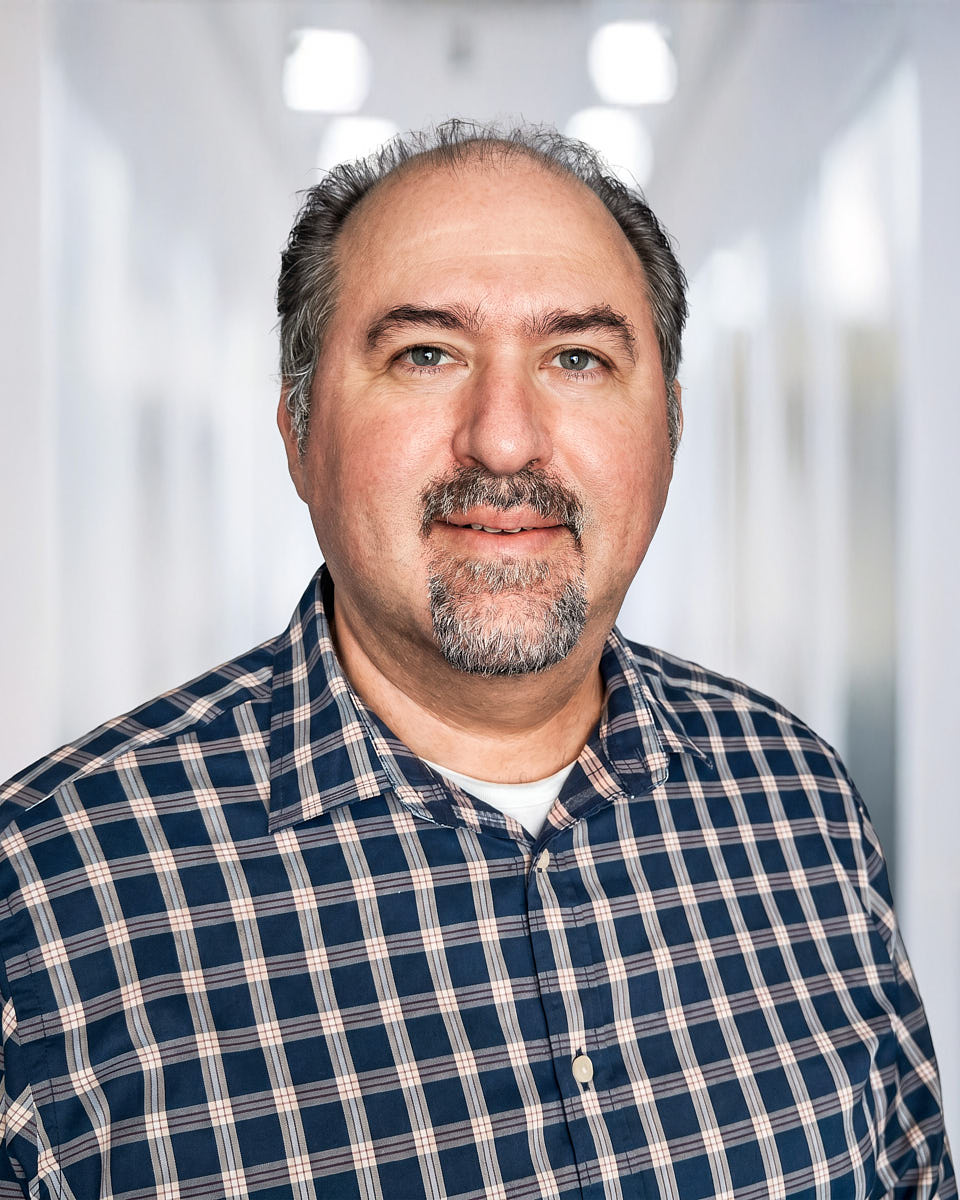 Natural executive headshot of a man in a plaid shirt with a softly blurred office setting.