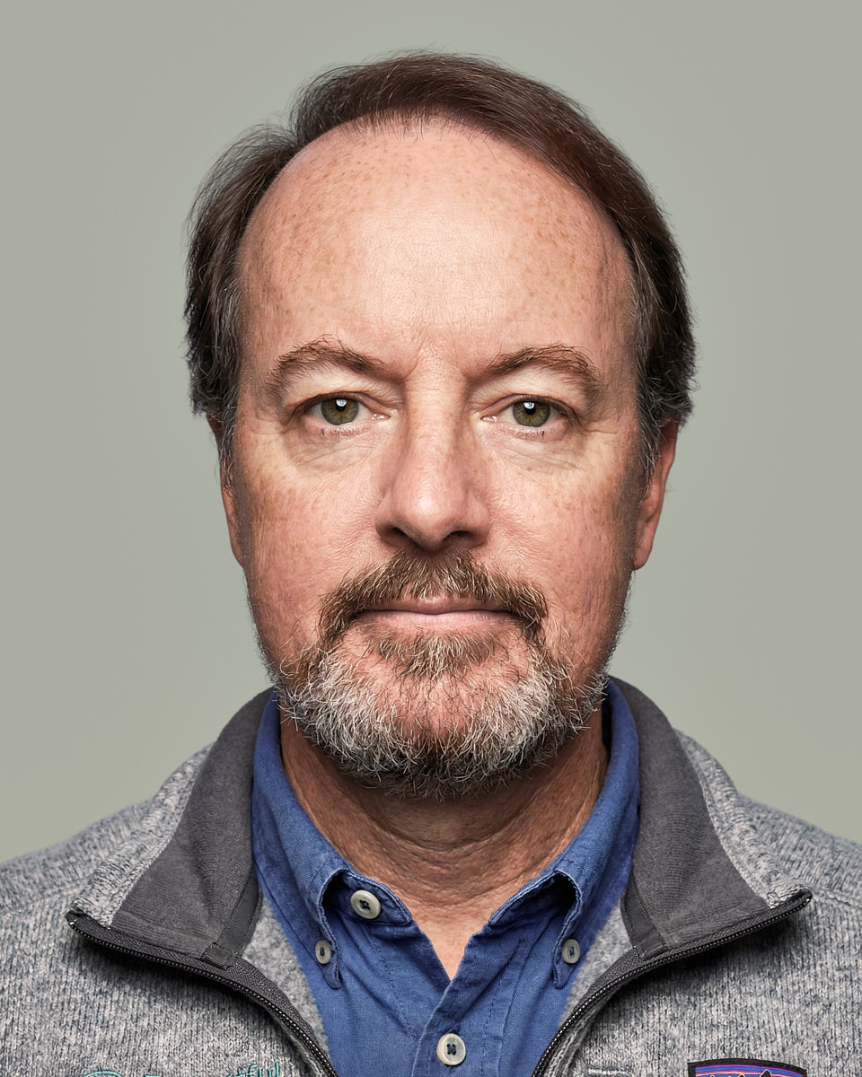 Consistent new hire headshot of a man in a gray sweater against a muted green background.