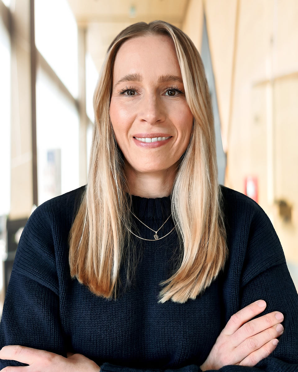 Remote team headshot of a blonde woman in a black top with a warm office-style background.